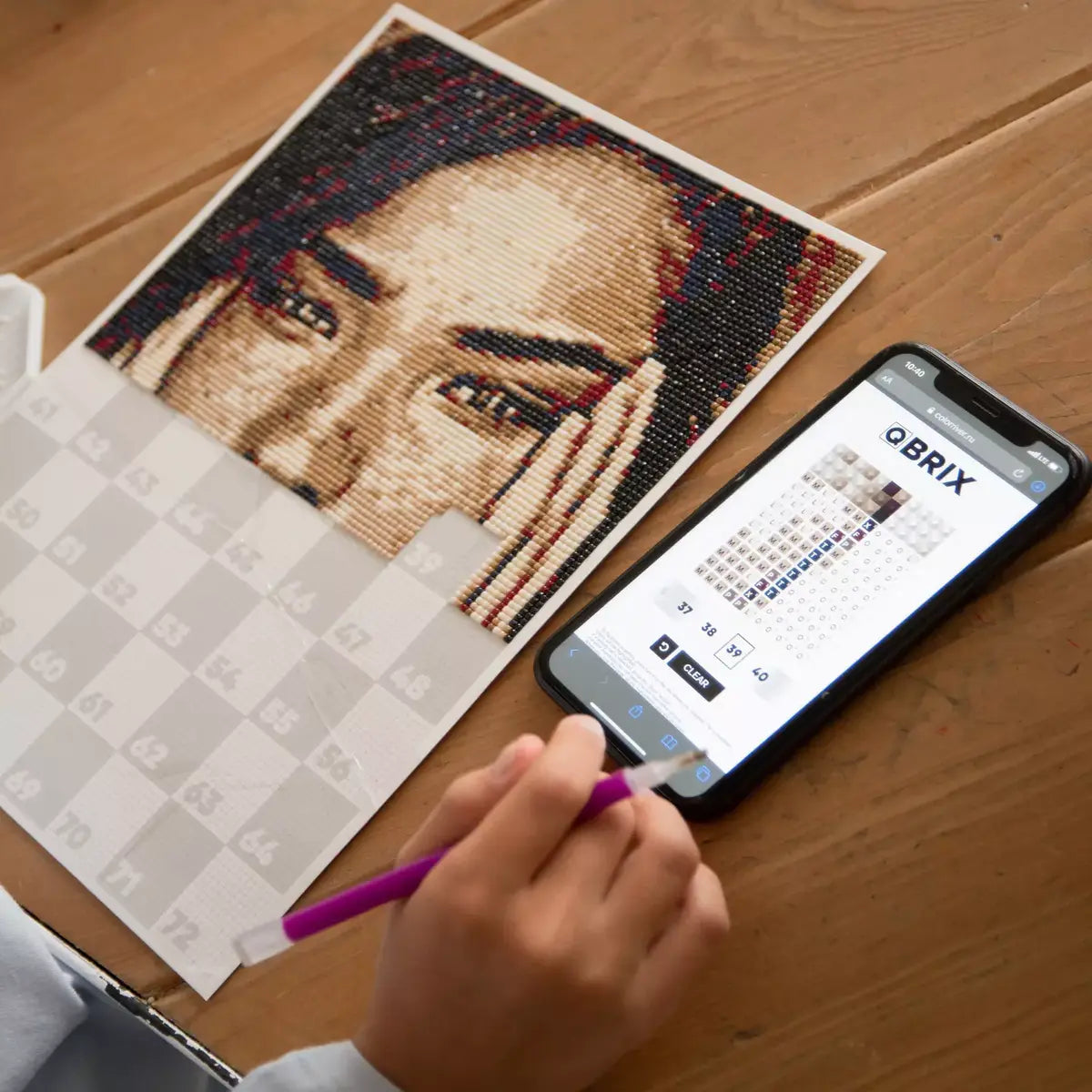 Person assembling a mosaic art piece with a smartphone displaying QBrix app on a wooden table.