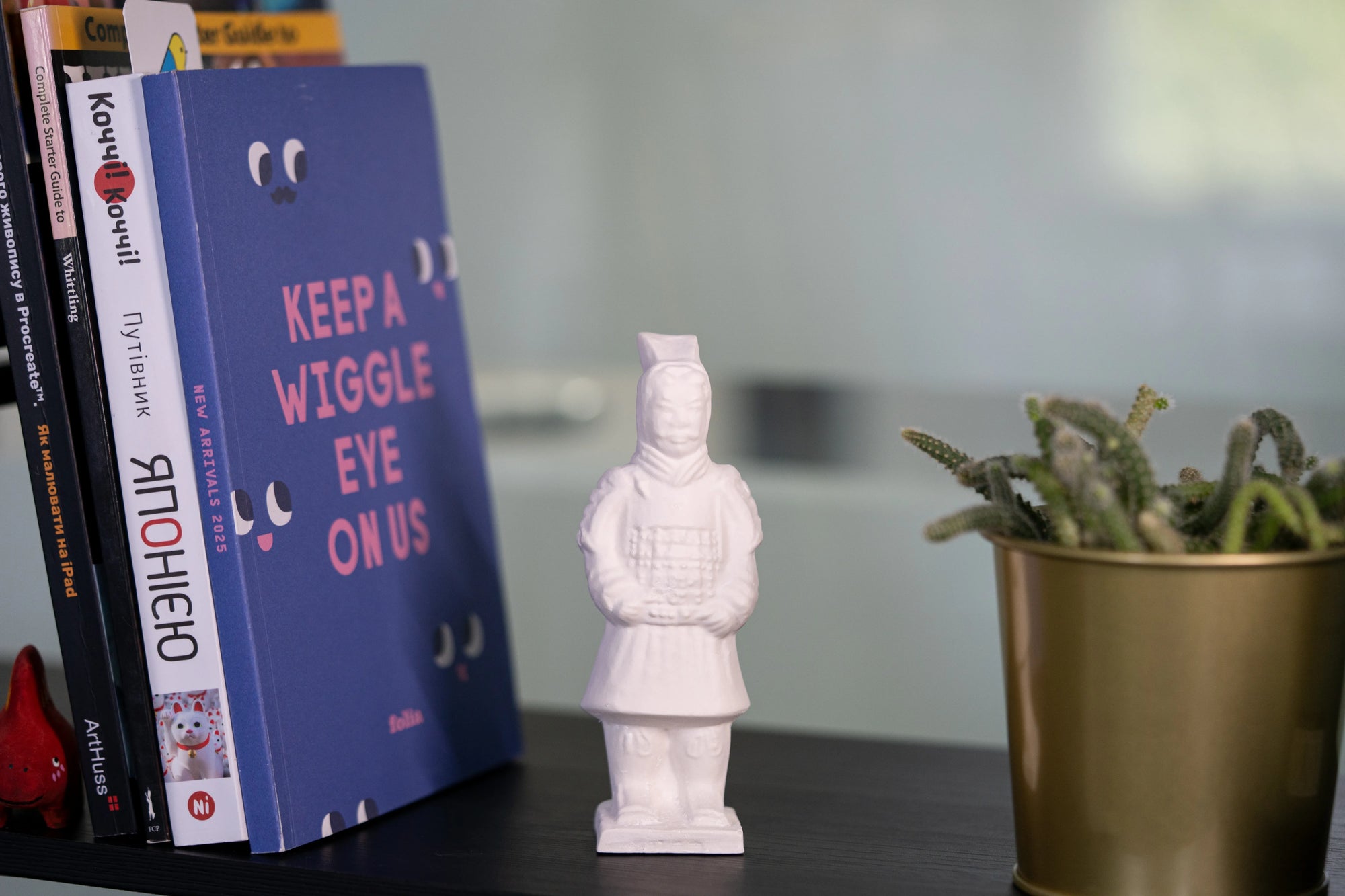 White warrior figurine next to a blue book titled 'Keep A Wiggle Eye On Us' on a shelf with books and a plant.